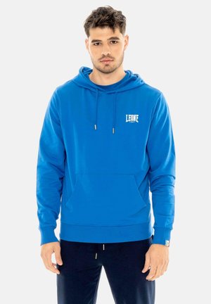 Joven con pelo corto y rizado, vistiendo una sudadera con capucha azul brillante con bolsillo delantero y pantalones negros, de pie frente a un fondo liso.
