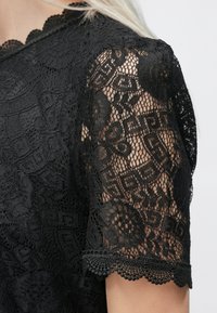 Μαύρη δαντελένια μπλούζα με λεπτομερείς λουλουδάτους σχεδιασμούς, κωνικό ντεκολτέ και κοντά μανίκια. Η υφή είναι πολύπλοκη με δι�άφανο αποτέλεσμα.