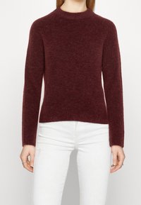 Pull en maille côtelée bordeaux avec col rond et manches longues. Le tissu semble doux et texturé, épousant étroitement le corps.