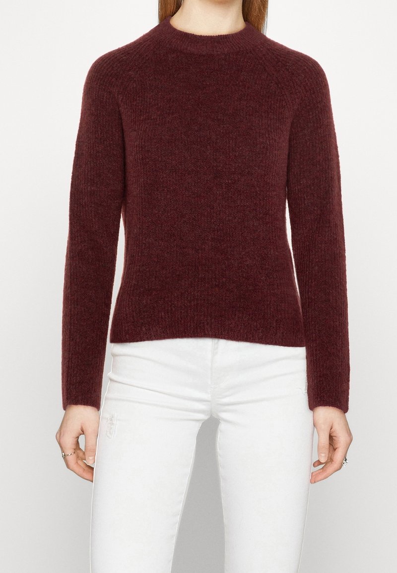 Pull en maille côtelée bordeaux avec col rond et manches longues. Le tissu semble doux et texturé, épousant étroitement le corps.