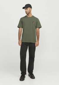 Camiseta de algodón verde oliva de manga corta, con un pequeño logo impreso en negro y rojo en el pecho izquierdo. Combinada con pantalones y zapatos negros.