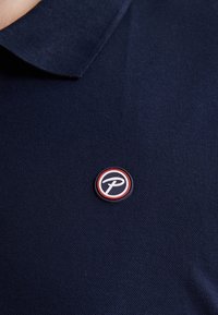 Marineblauwe polo met een textuurstof en een ronde patch met een gestileerd logo in rode en witte accenten aan de linkerkant.