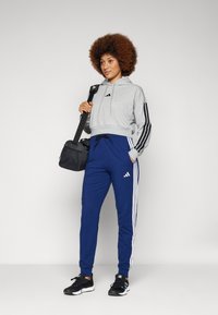 Grå kort hoodie med svarta ränder, tillsammans med blå träningsoverallsbyxor med vita sidostråk. Svarta sneakers och en svart axelväska.