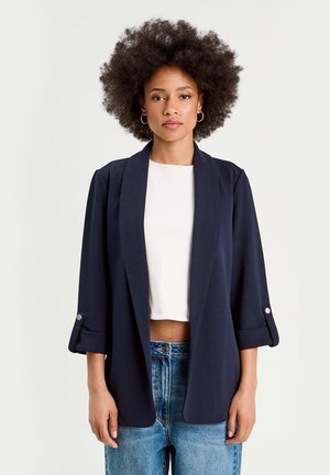 Blazer blu navy con collo scialle e maniche con risvolto. Realizzato in tessuto morbido, presenta una vestibilità rilassata e un design minimalista.