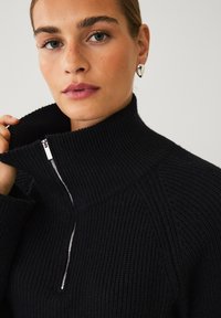 Sort strikket pullover med høj krave og sølv lynlåsdetalje. Har ribbet tekstur og lange ærmer, som viser et nærbillede.