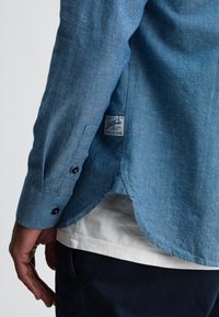 Blauwe chambray overhemd met lange mouwen en knoopmanchetten, afgeronde zoom en een label nabij de zijnaad. Gekocht over een wit t-shirt.