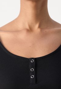 Gros plan du cou et de la clavicule d'une personne portant un haut côtelé noir avec trois boutons-pression argentés au centre devant.