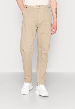 Homme portant un pantalon décontracté beige avec des poches avant et des baskets blanches, debout devant un fond gris clair uni.