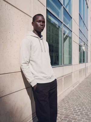 Jeune homme en sweat à capuche gris clair et pantalon noir, debout contre un mur en pierre beige avec de grandes fenêtres en verre dans une rue urbaine ensoleillée.