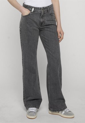 Person iført høytlivs, løstsittende mørkegrå jeans med hånden i lommen, kombinert med grå og hvite joggesko mot en ensfarget bakgrunn.