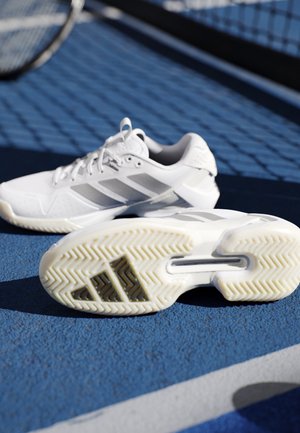 Witte sportschoenen met grijze strepen op een blauw tennisveld, één schoen toont de zool met textuurprofiel en logo.