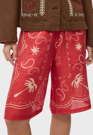 Røde shorts med hvide paisley- og palmetræsprint båret sammen med en brun jakke med broderede blomster- og løvmønstre.