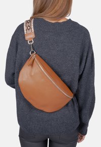 Sac en cuir marron en bandoulière avec une forme incurvée et une fermeture éclair argentée. La bandoulière présente un motif tissé en crème et marron. Porté sur un pull gris.