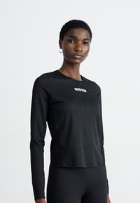 Vrouw in een zwart aansluitend longsleeve shirt en zwarte broek, staand tegen een effen witte achtergrond.