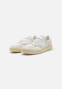 Weiße Leder-Sneaker mit perforierter Zehe, flacher Schnürdesign und subtilen grauen Akzenten. Verfügt über eine cremefarbene Gummisohle ohne sichtbares Branding.