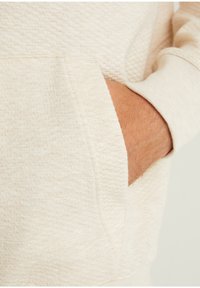 Hoodie beige clair avec un tissu texturé, une poche avant et des poignets côtelés. Le matériau semble doux avec un motif subtil.