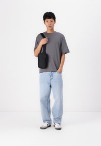 Tricou gri cu mânecă scurtă, blugi maro deschis cu croială largă și geantă de umăr neagră; stând pe un fundal alb simplu.