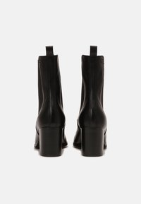 Par de botas de cuero negras hasta el tobillo con tacones cuadrados, vistas desde la parte de atrás sobre un fondo blanco liso.