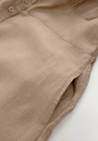 Robe en lin beige avec patte de boutonnage, poche latérale et détails froncés sur l'épaule pour une coupe décontractée et une texture légère.