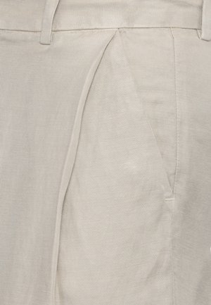 Primo piano di pantaloni in tessuto beige che mostrano la vita, i passanti per la cintura e una tasca laterale con dettagli di cuciture visibili.