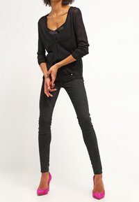 Svart transparent cardigan med långa ärmar, lager över en svart topp. Parat med figurnära svarta jeans och klarröda högklackade skor.