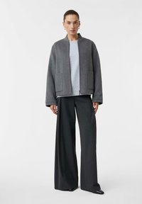 Frau trägt eine graue Zip-Jacke, einen hellgrauen Pullover, weite dunkelgraue Hosen und schwarze spitze Schuhe und steht vor einem weißen Hintergrund.