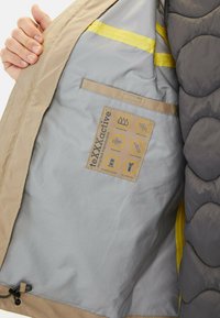 Beige og grå jakke indvendigt med et glat, gråt stof; har gule detaljer, et mærke med vandtætte og åndbare ikoner samt en snor.