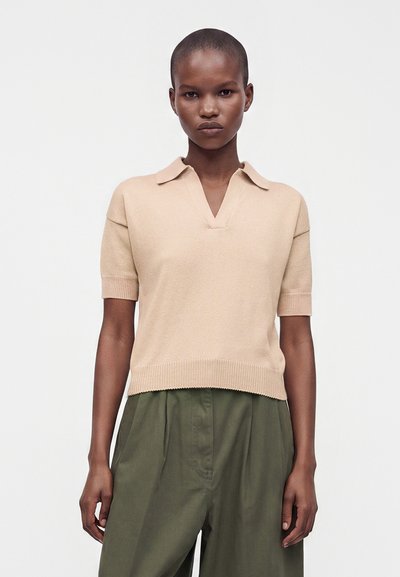 Femme aux cheveux courts portant un haut en maille beige à manches courtes avec col et un pantalon taille haute vert olive sur fond blanc uni.