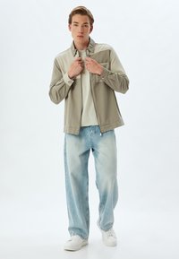 Lichte, twee-kleurige jas in beige en kaki, met een ritsluiting. Gepaard met vervaagde blauwe wijde jeans en witte sneakers.
