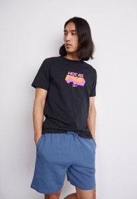 T-shirt en coton noir avec un texte graphique audacieux et coloré "BRÛLANT COMME F*%#!" sur la poitrine ; associé à un short bleu à cordon. 