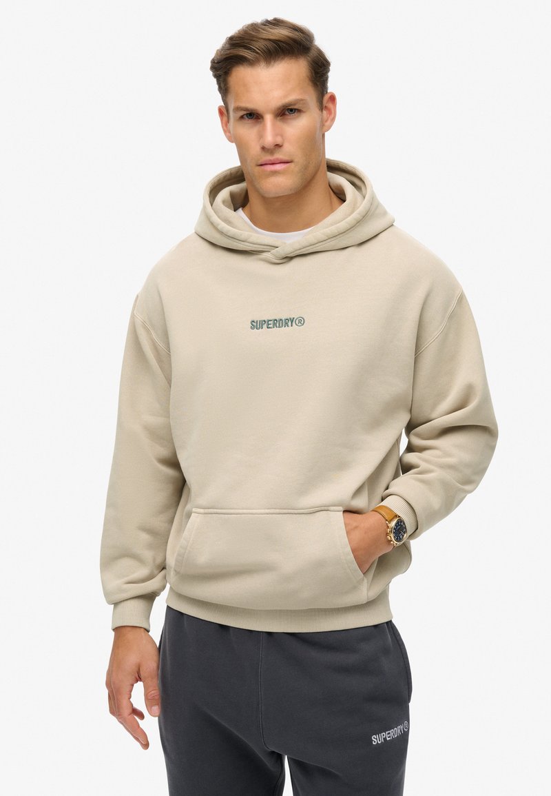 Sudadera con capucha beige y bolsillo frontal, con un logo bordado en verde "SUPERDRY". Textura suave, corte relajado, puños acanalados.