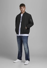 Crna bomber jakna s džepom na patentni zatvarač, nošena preko bijele majice, u kombinaciji s plavim trapericama i bijelim tenisicama. Glatki materijal, ležerni dizajn.