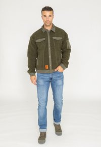 Oliwkowa polarowa kurtka z zapięciem na guziki, dwiema kieszeniami na piersi i ściągaczami na rękawach. Połączona z jasnoniebieskimi dżinsami i zielonymi butami.