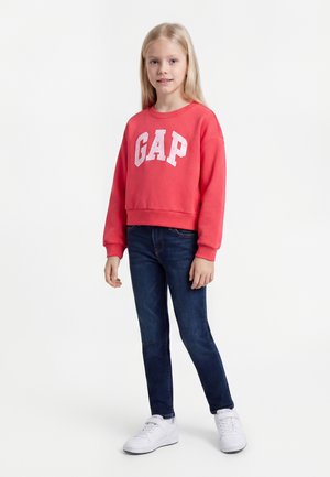 Νεαρή κοπέλα που φοράει κόκκινο φούτερ GAP, σκούρα μπλε τζιν και λευκά αθλητικά παπούτσια σε απλό λευκό φόντο.