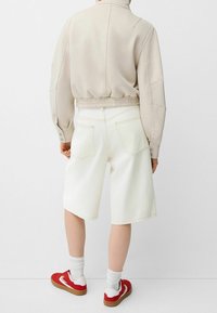 Light beige cropped jacket with elasticated waistband, paired with white denim shorts. Red sneakers with white and tan accents complete the outfit.