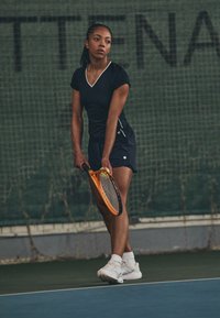 Camiseta atlética negra con cuello en V y detalles en blanco, combinada con pantalones cortos negros. El jugador sostiene una raqueta de tenis naranja en una cancha.