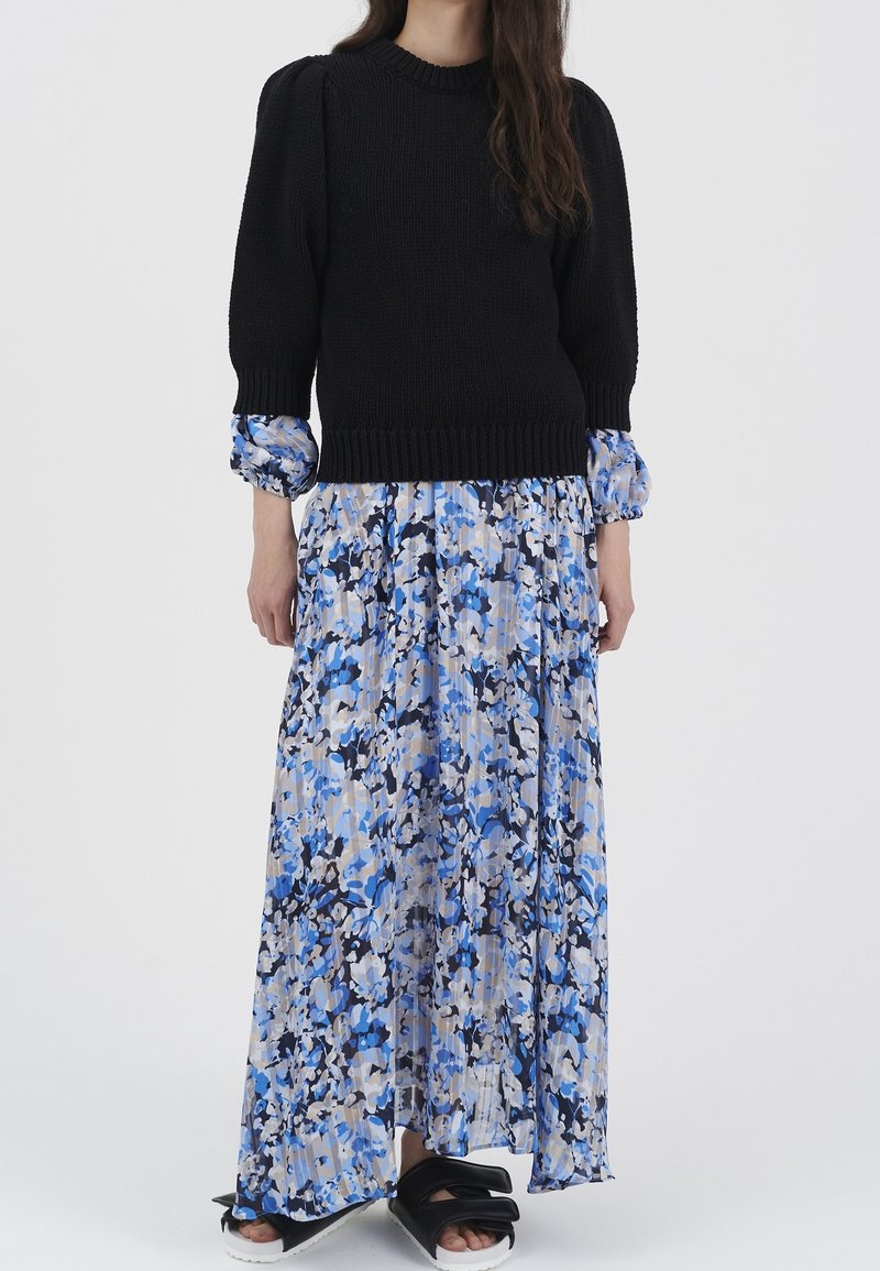 Femme portant un pull en maille noir sur une longue jupe plissée fleurie bleu, blanc et noir avec des sandales noires à bout ouvert sur fond blanc.