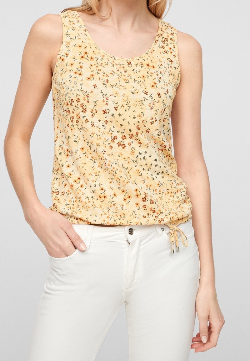 Femme portant un haut jaune à fleurs sans manches avec un lien réglable sur le côté et un pantalon blanc, main reposant dans la poche.