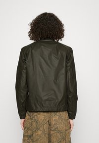 Mörkgrön bomberjacka med en slät, glänsande yta, elastisk midja och avsmalnande ärmar; bärs över byxor med camouflage-mönster.