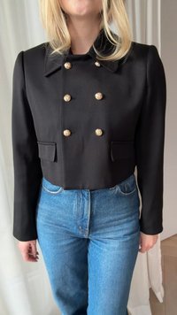 Donna che indossa una giacca corta doppiopetto nera con bottoni dorati e jeans azzurri a vita alta su uno sfondo neutro.