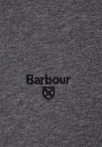 Tmavě šedá látka s černě vyšitým textem "Barbour" a emblémem uprostřed povrchu.