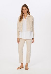 Beige, verkürzte Denimjacke mit goldenen Details, kombiniert mit einer weißen gestreiften Bluse, beigen Hosen und goldenen Sandalen.
