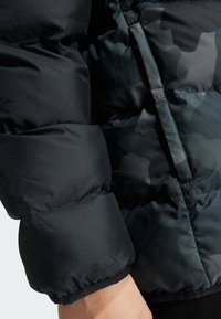 Veste matelassée noire avec une finition brillante, présentant un motif camouflage sombre subtil, des sections matelassées et des poignets élastiques.