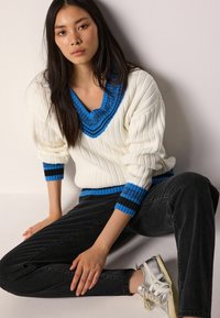 Weißer Strickpullover mit blauen und schwarzen Akzenten, tiefem V-Ausschnitt, gerippter Textur, kombiniert mit schwarzen Jeans und silbernen Sneakers.