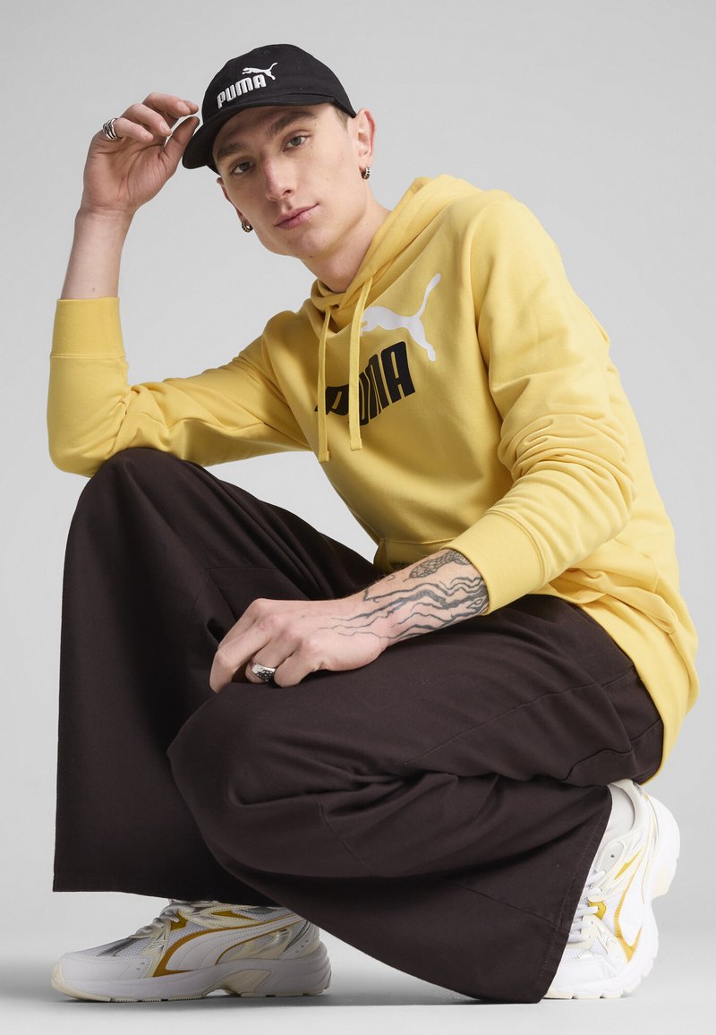 Felpa gialla con cappuccio e logo nero, pantaloni marroni larghi e scarpe da ginnastica bianche con dettagli gialli, accompagnati da un cappellino nero.