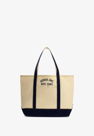Beżowa torba tote z kanwy z granatowymi akcentami i uchwytami. Na przodzie haftowany napis "GUESS INC. EST. 1981". Ma wyraźny kształt.