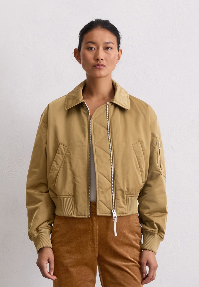 Woman wearing a tan bomber jacket over a white top and brown pants, standing against a plain light gray background.