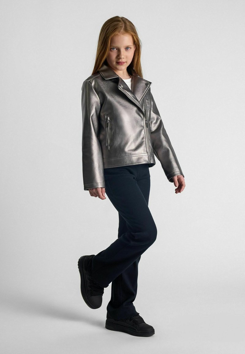 Veste en simili cuir argent métallique avec col à revers, poches zippées et manches longues; associée à un pantalon sombre et des baskets noires.