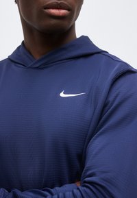 Κοντινό πλάνο ενός ατόμου που φορά γαλάζιο φούτερ με κουκούλα Nike με σταυρωμένα χέρια, δείχνοντας το λευκό λογότυπο Nike swoosh στο στήθος.