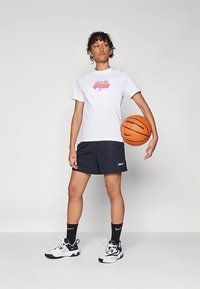 Balts T-krekls ar violeta un rozā grafisku tekstu, melnas šortus, baltas un melnas sporta kurpes un melnas zeķes; tur rok�ās basketbola bumbu.